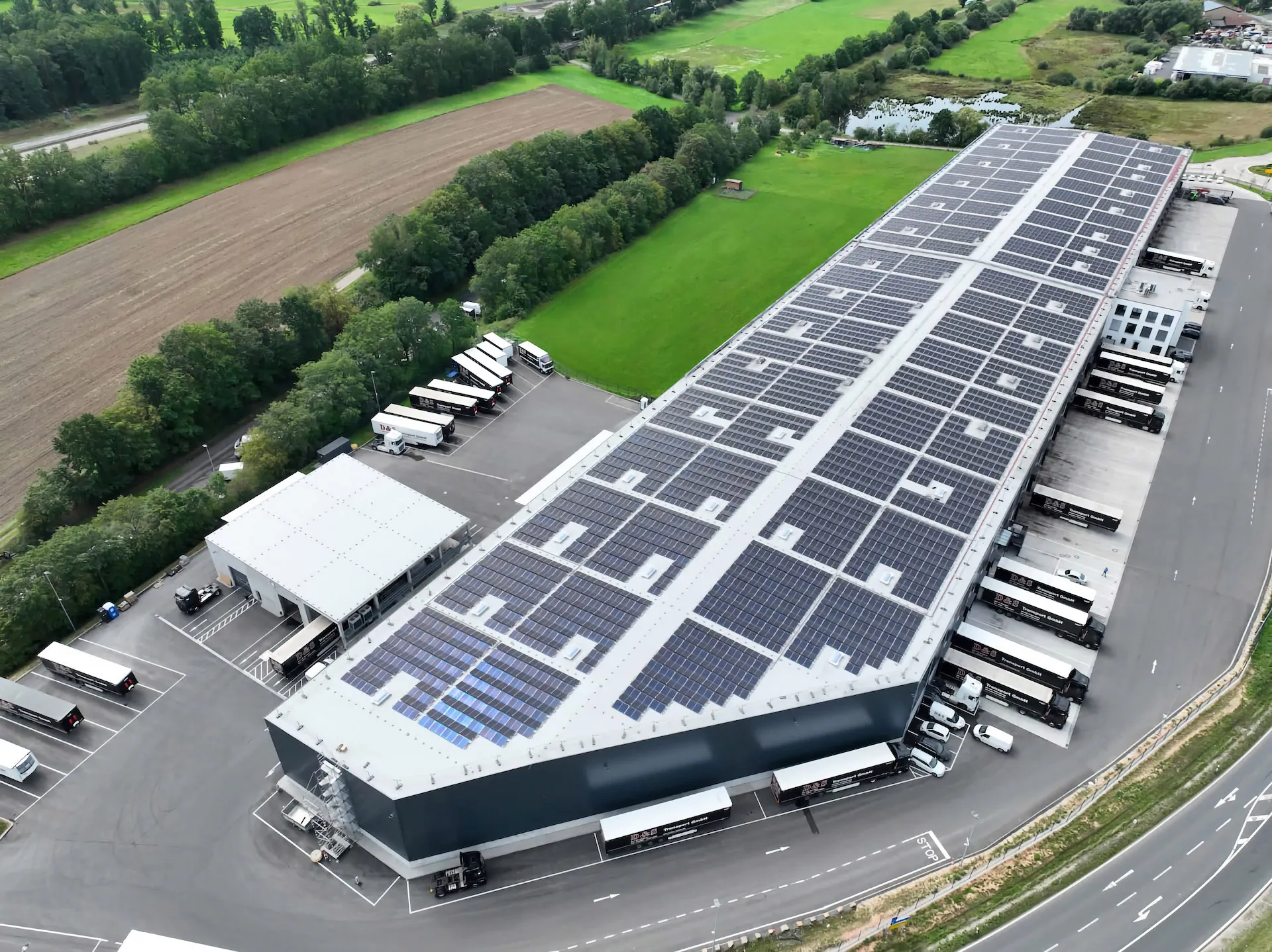 Aerial view of a large warehouse with solar panels on roof and trucks parked outside.