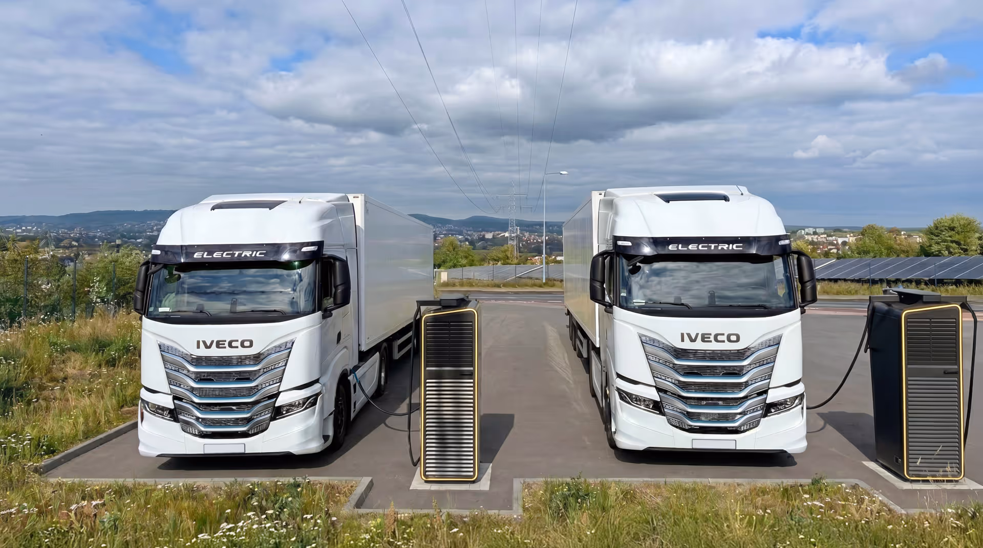 Zwei weiße Elektro-Lkw laden an ESG Ladestationen auf einem Parkplatz bei bewölktem Himmel.