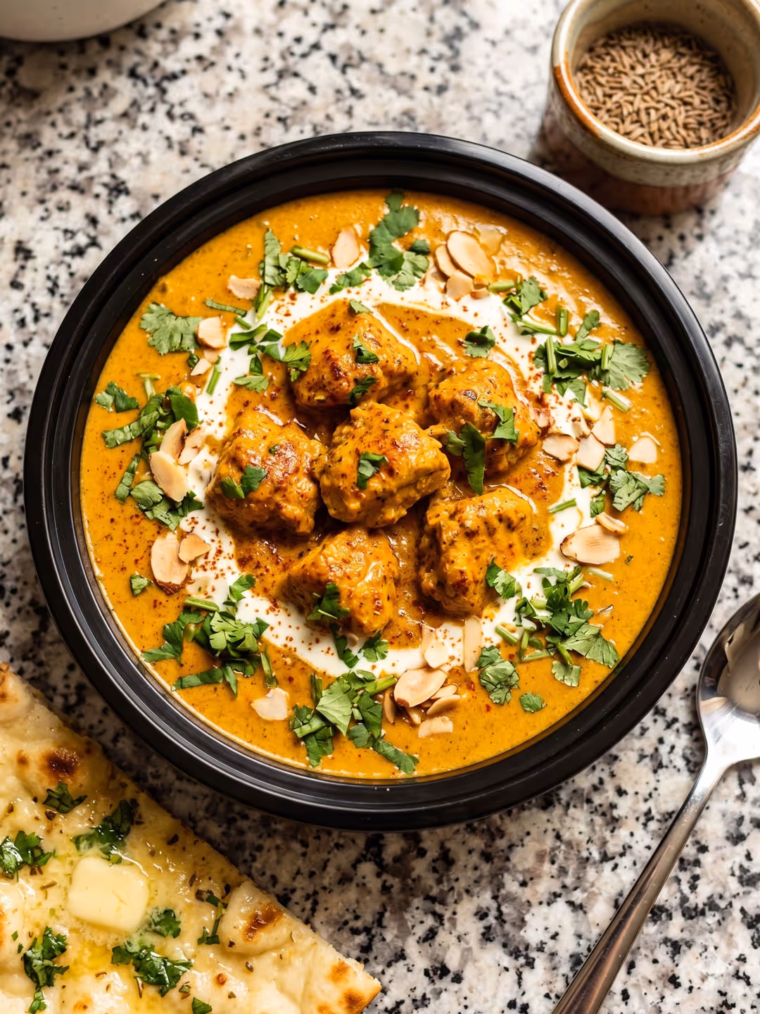 Spiced curry with chunks, garnished with herbs and almonds, served with butter naan bread and spoon.