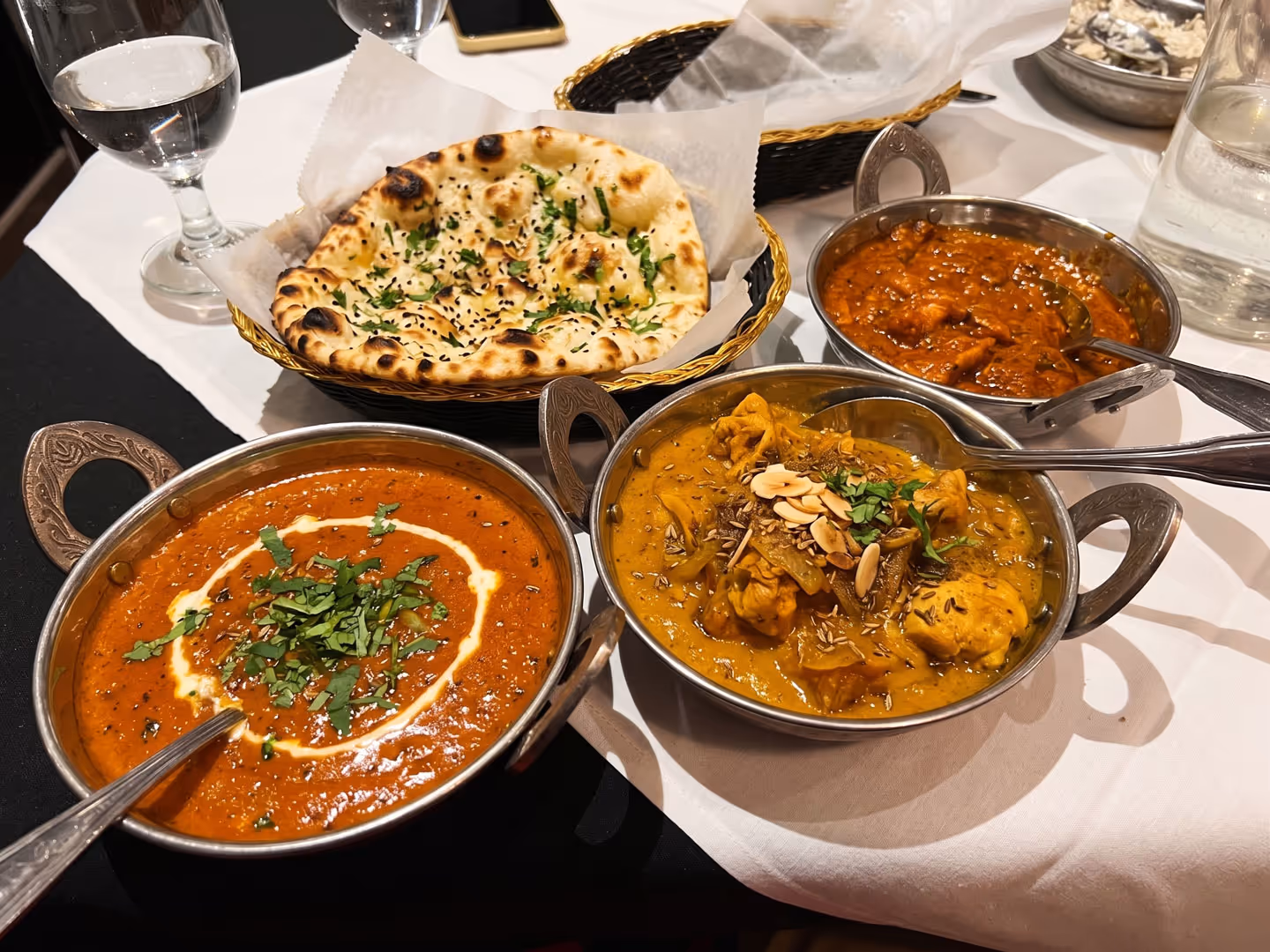 Three Indian curry dishes and a basket of naan bread served on a white tablecloth with glasses of water.