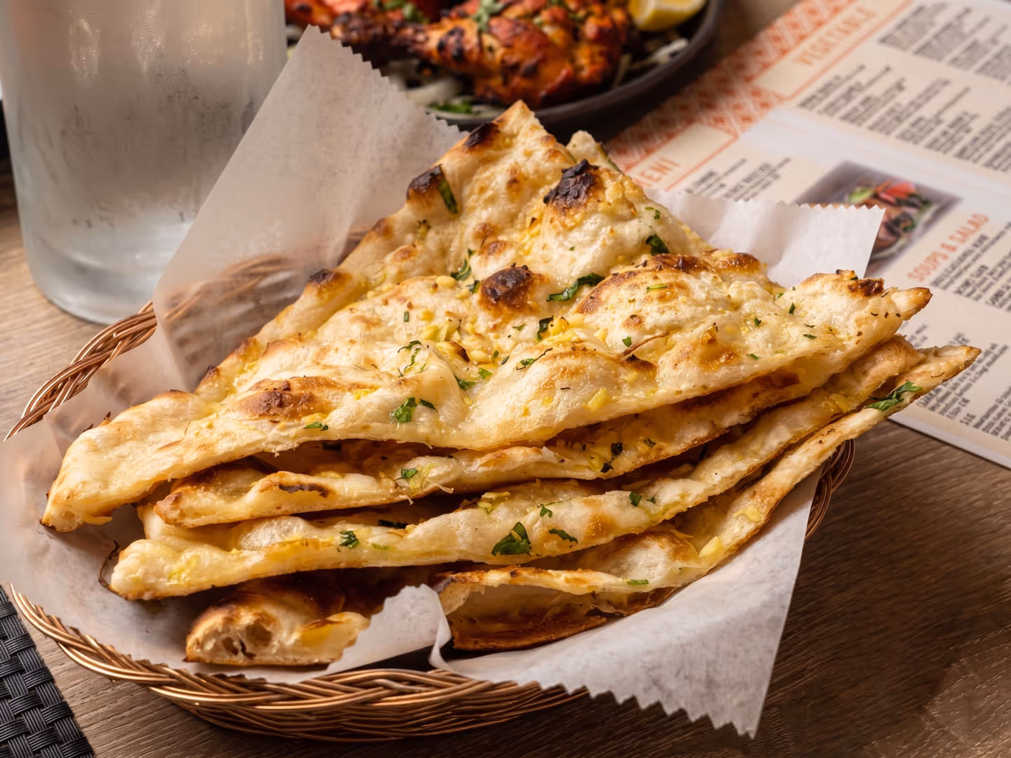 Basket of garlic naan bread with herbs on parchment paper on a wooden table near a menu and glass.