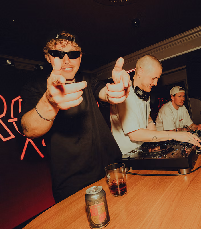 En person i solglasögon pekar med fingrarna mot kameran medan två andra står vid DJ-utrustning.