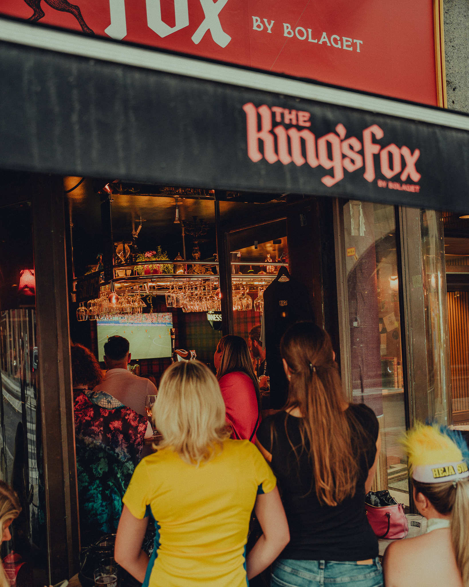 People watching a soccer game inside a pub named The King's Fox from outside the entrance.