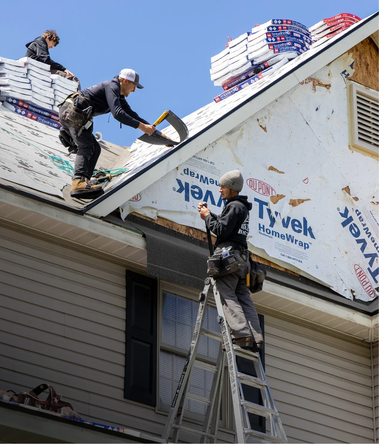 Two workers install roofing materials on a house, one on the roof and one on a ladder, with stacked shingles visible above them.