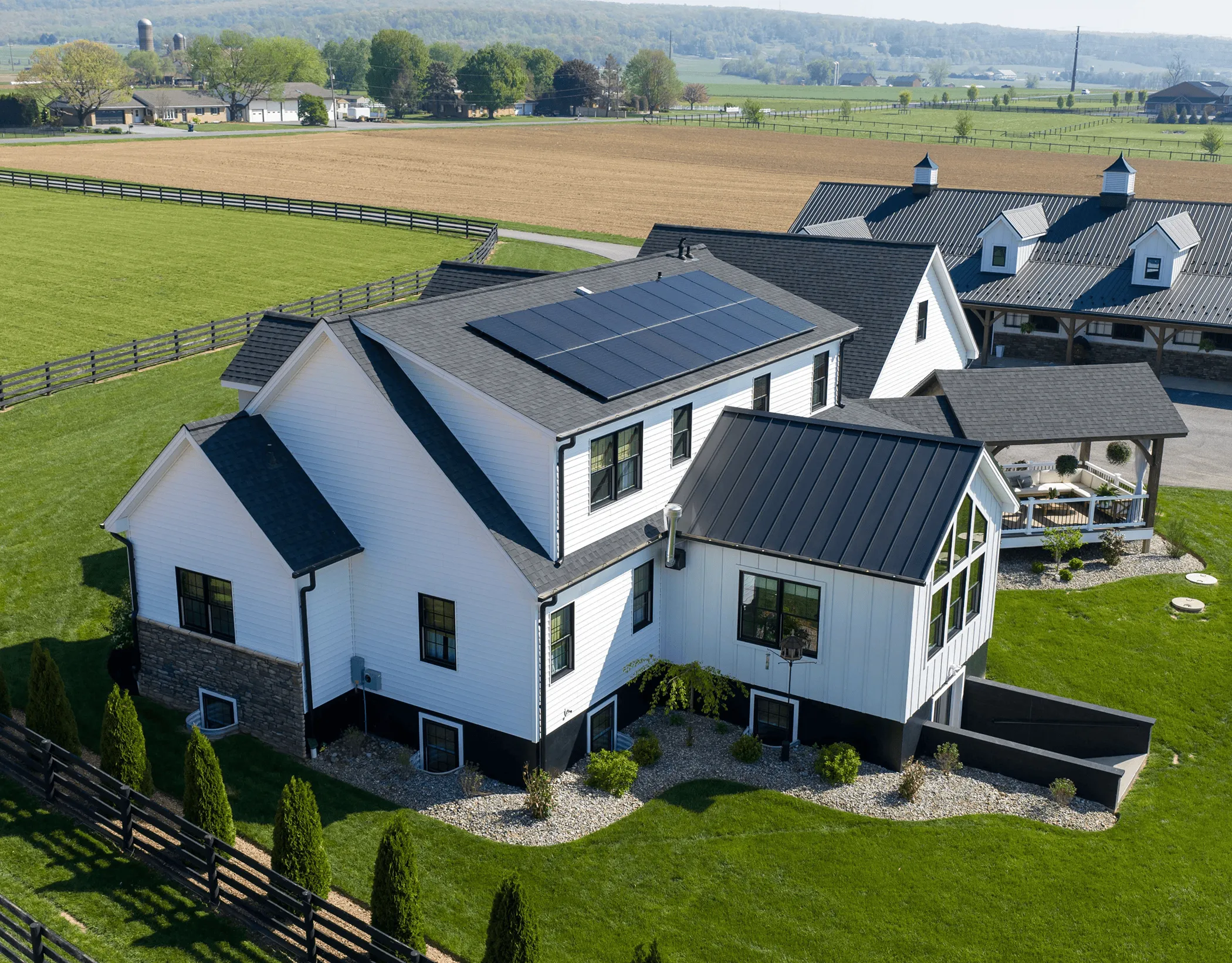 A modern white farmhouse with solar panels on the roof, surrounded by green fields and landscaping.