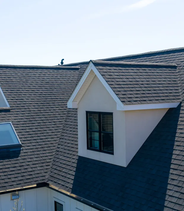 A close-up view of a house roof with dark shingles, a dormer window, and skylights under a clear sky.