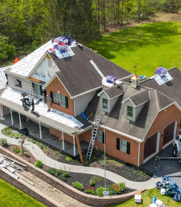 Workers are installing new roof shingles on a two-story brick house using ladders and roofing materials.
