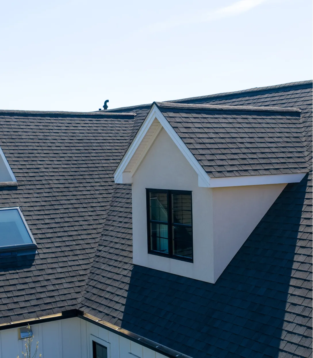 A close-up view of a house roof with dark shingles, a dormer window, and skylights under a clear sky.
