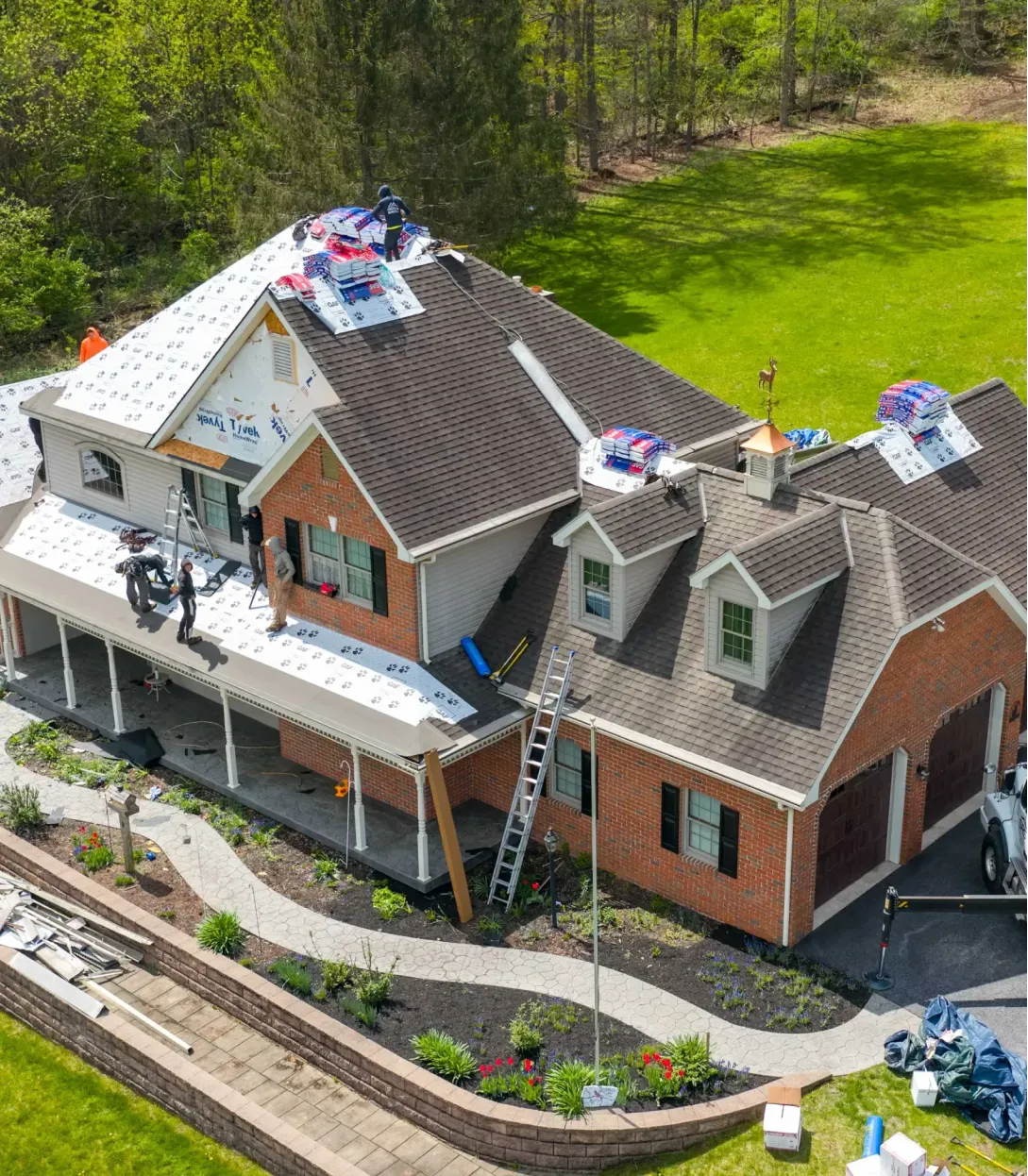 Workers are installing new roof shingles on a two-story brick house using ladders and roofing materials.