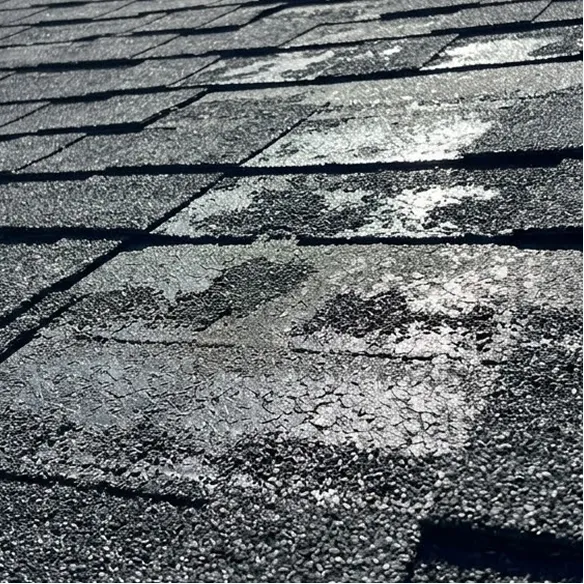 Close-up of asphalt roof shingles with several areas showing visible wear, granule loss, and patches where shingles appear damaged or deteriorated.