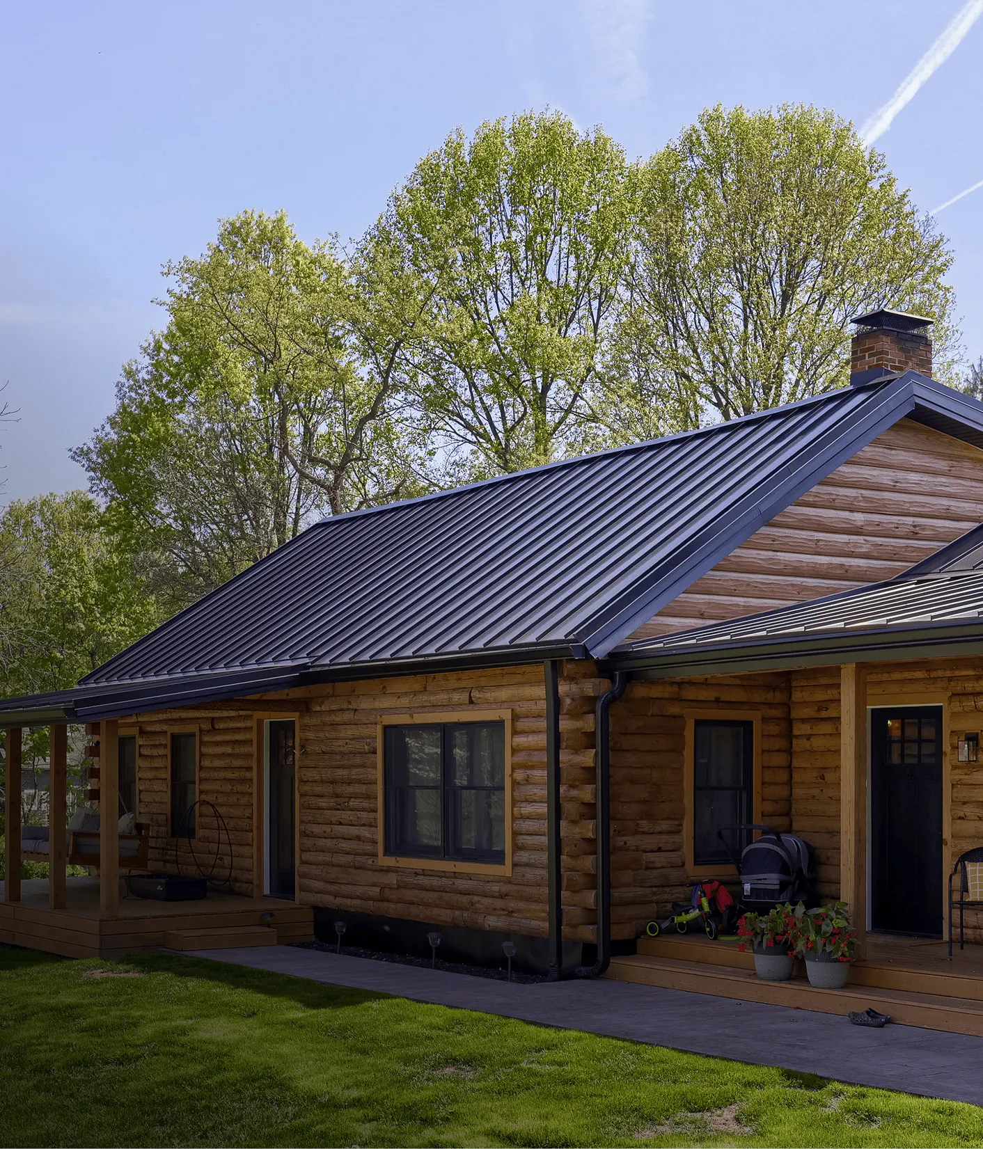 A single-story log cabin with a metal roof, covered porch, and potted plants sits on a green lawn.