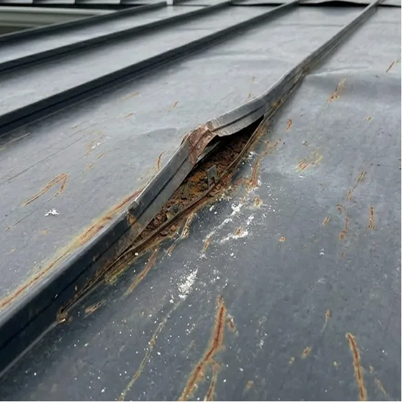 Close-up of a metal roof with a bent and damaged seam, showing signs of rust and weathering.