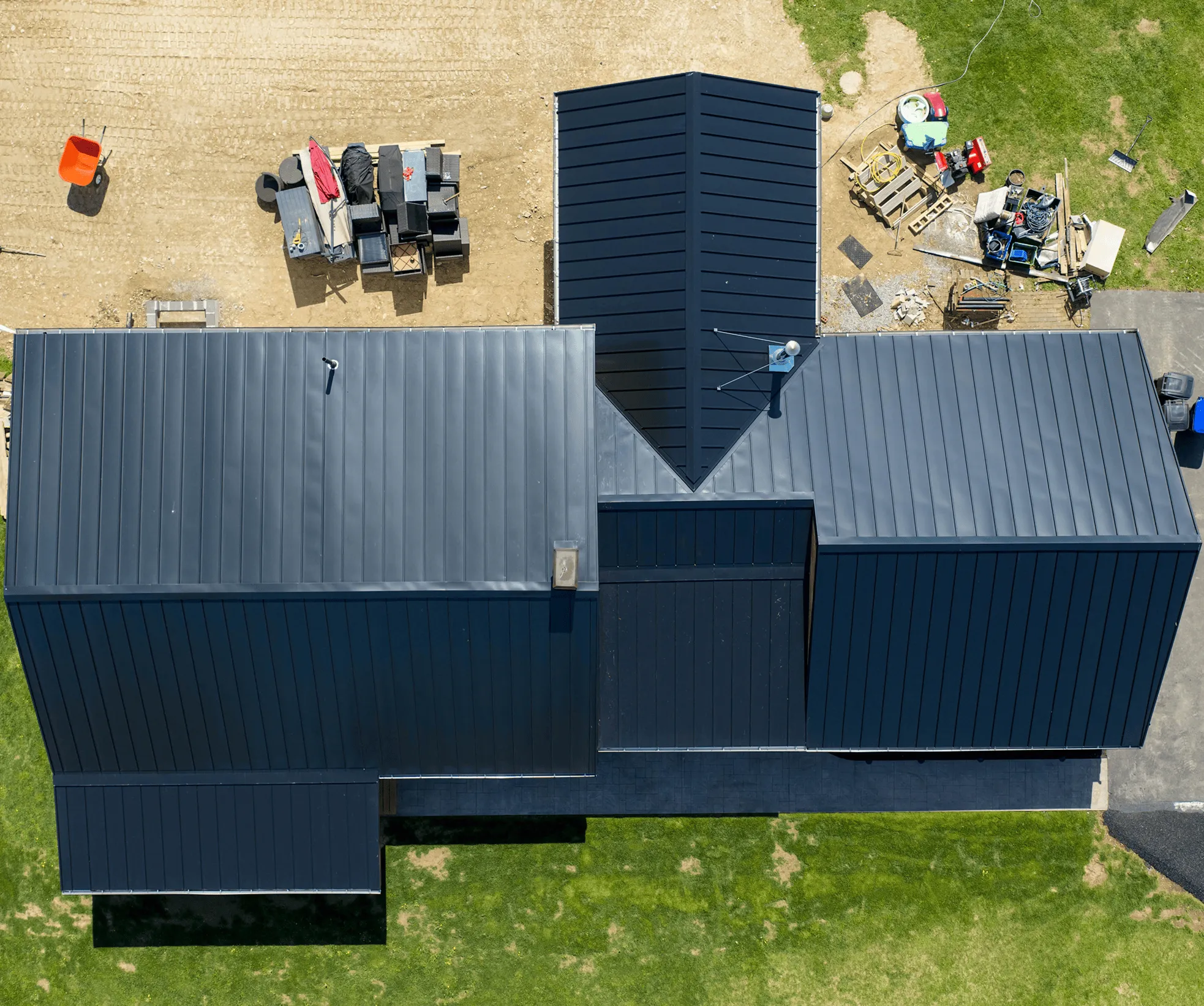 Aerial view of a modern black metal-roofed building with construction materials and equipment scattered on the surrounding dirt and grass.