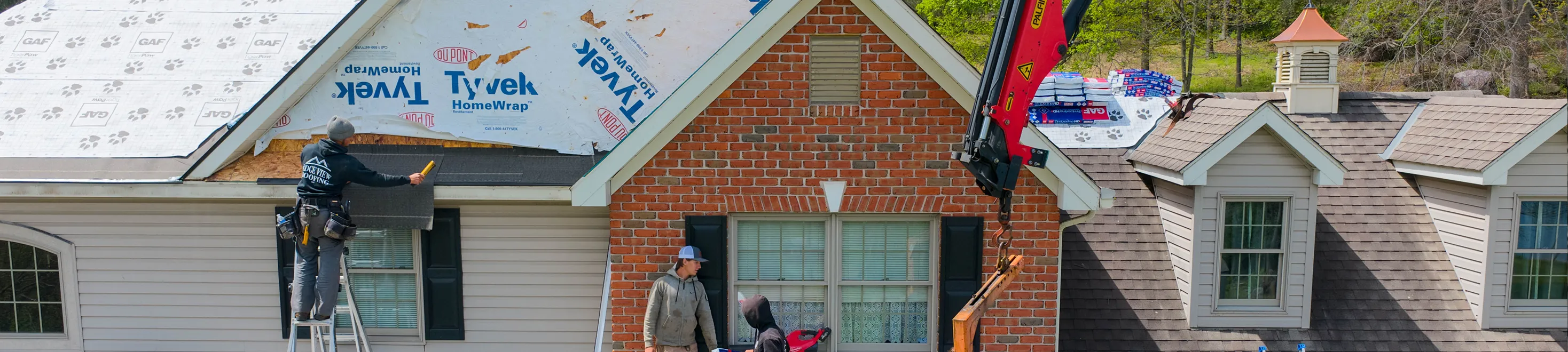 Two workers install new roofing materials on a house; one stands on a ladder, while the other is on the ground near construction equipment.