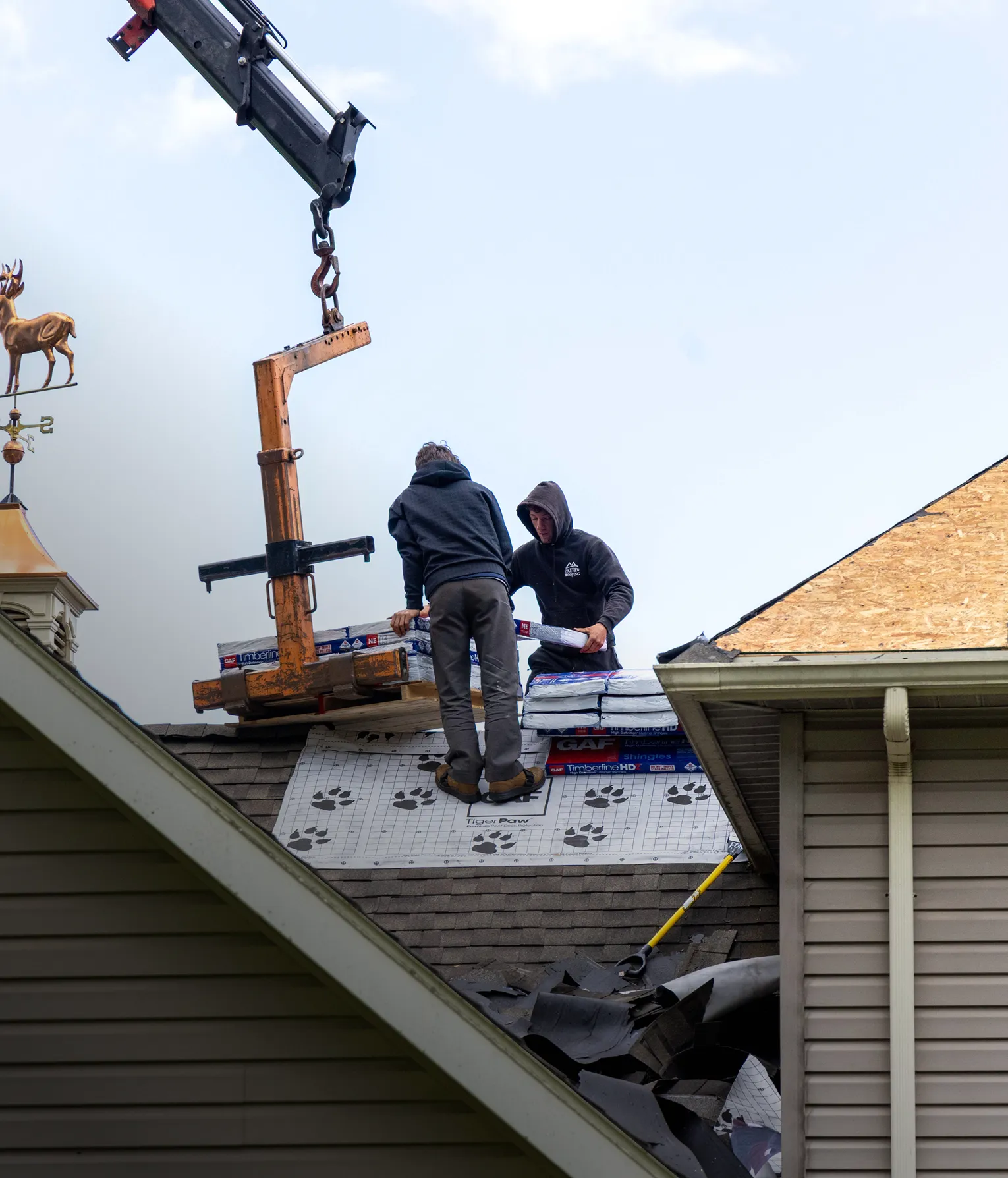Two workers on a roof unload shingles using a crane, with roofing materials and debris visible around them on a partially finished roof.