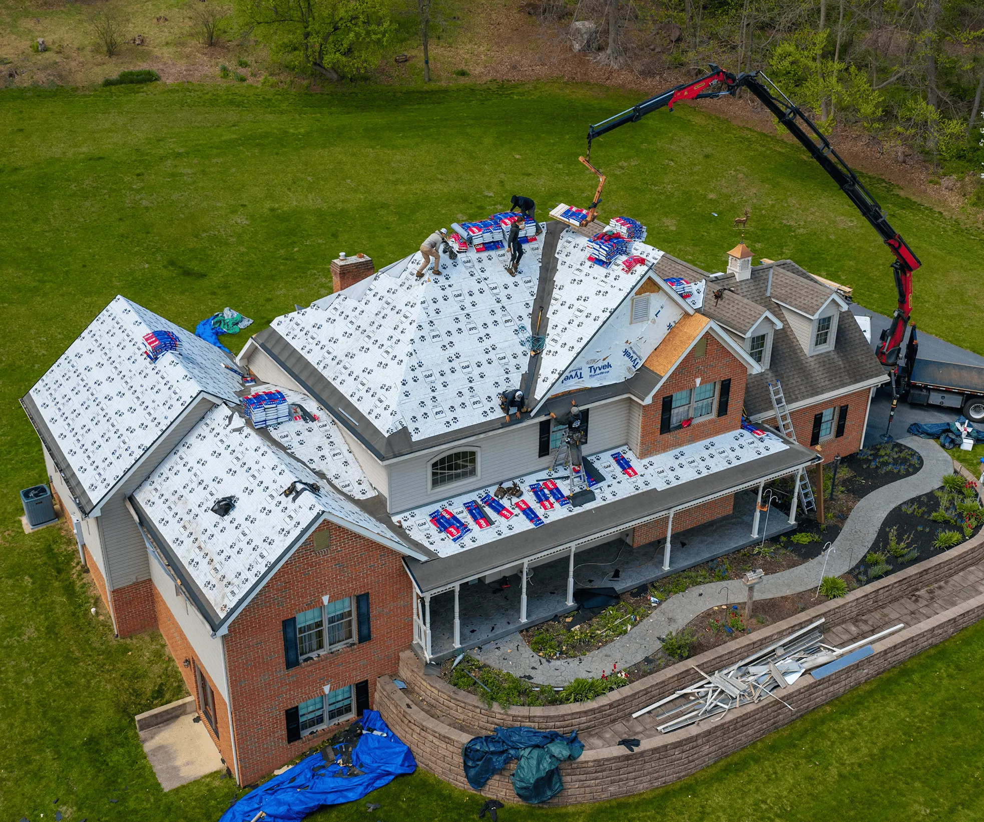 Aerial view of workers installing new roofing materials on a large house, with tools, supplies, and a crane truck visible on the property.