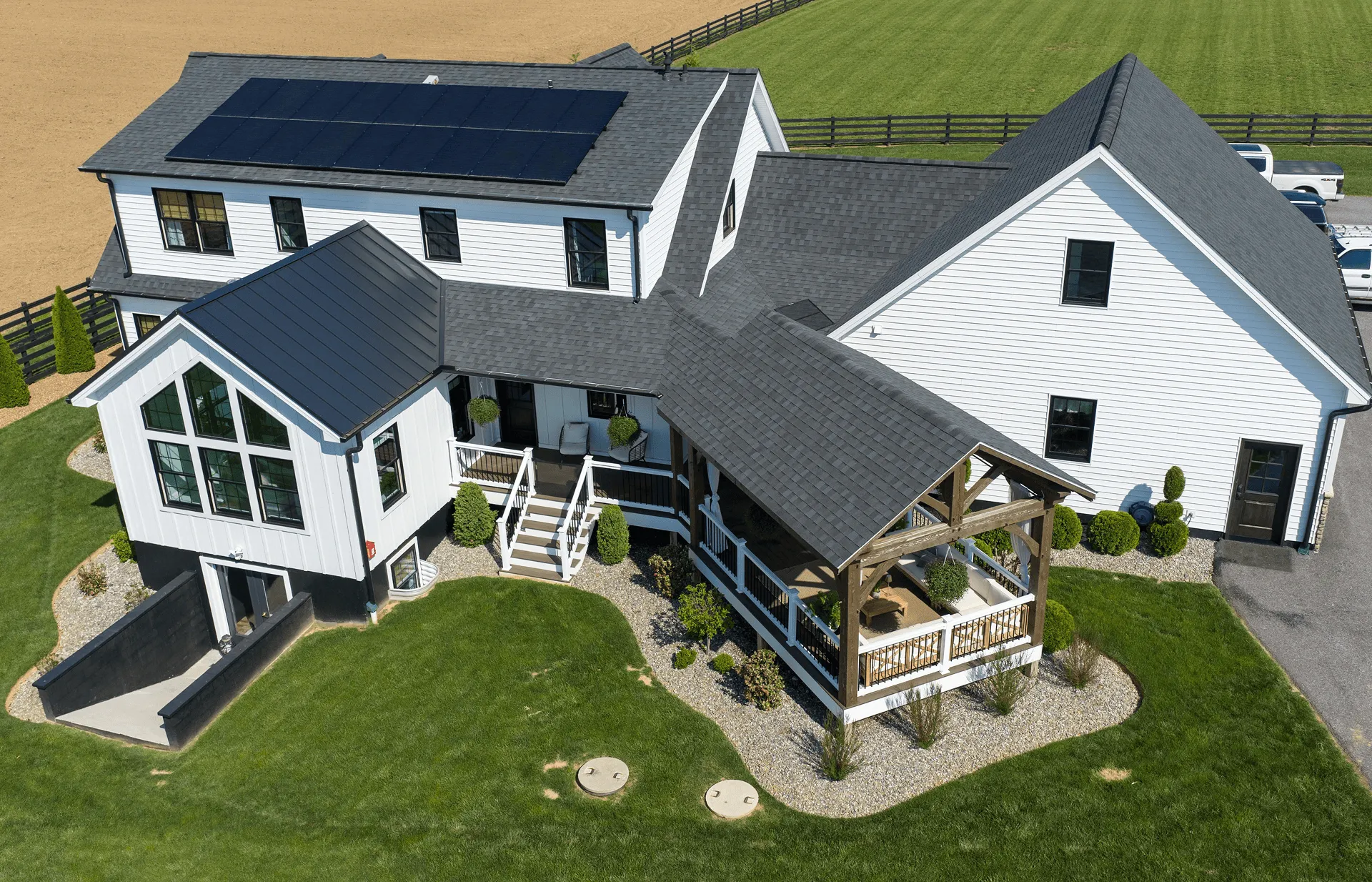 Aerial view of a modern farmhouse with white siding, black roof, solar panels, large porch, and surrounding green lawn.