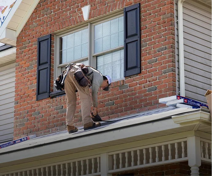 A team member wearing work gear is repairing shingles on the roof of a brick house beneath a window with black shutters.