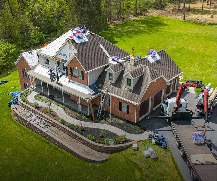 Aerial view of workers installing new shingles on the roof of a two-story brick house using ladders and equipment, with materials and a truck on the lawn.