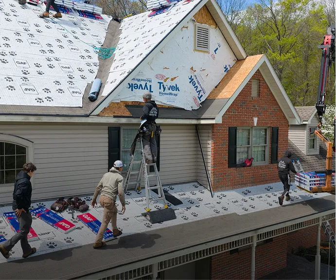 Several workers are installing roofing materials on a house, some on the roof and others on ladders, with tools and roofing supplies visible around them.