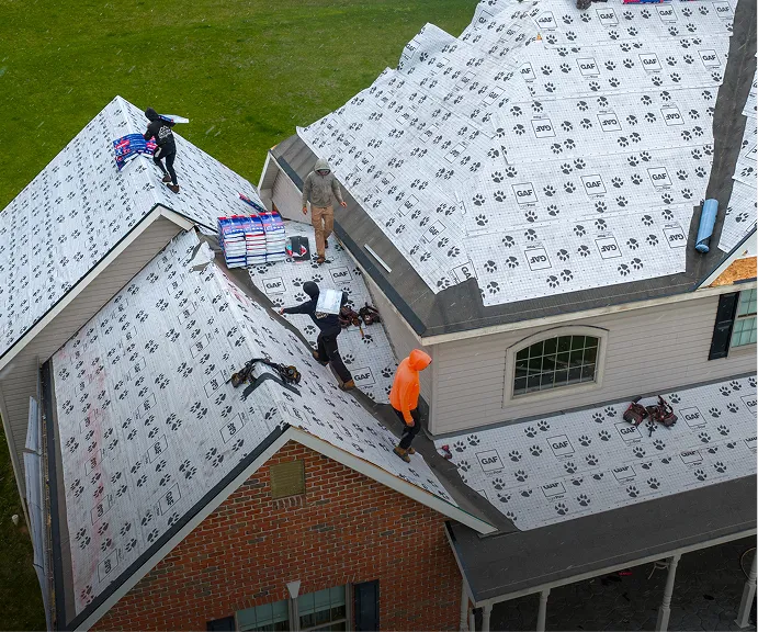 Four workers install roofing underlayment on a house roof, with tools and materials visible; the roof is steep and partially covered in synthetic underlayment sheets.