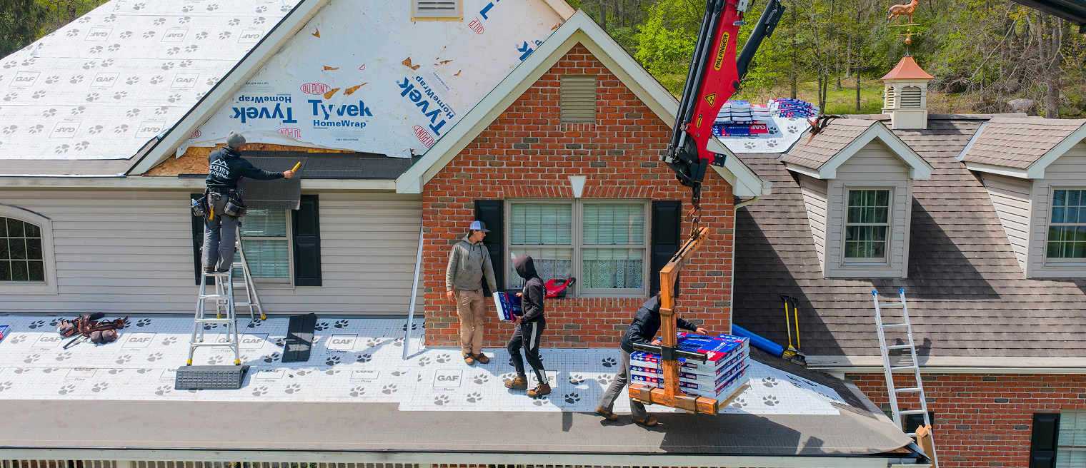 Two workers install new roofing materials on a house; one stands on a ladder, while the other is on the ground near construction equipment.