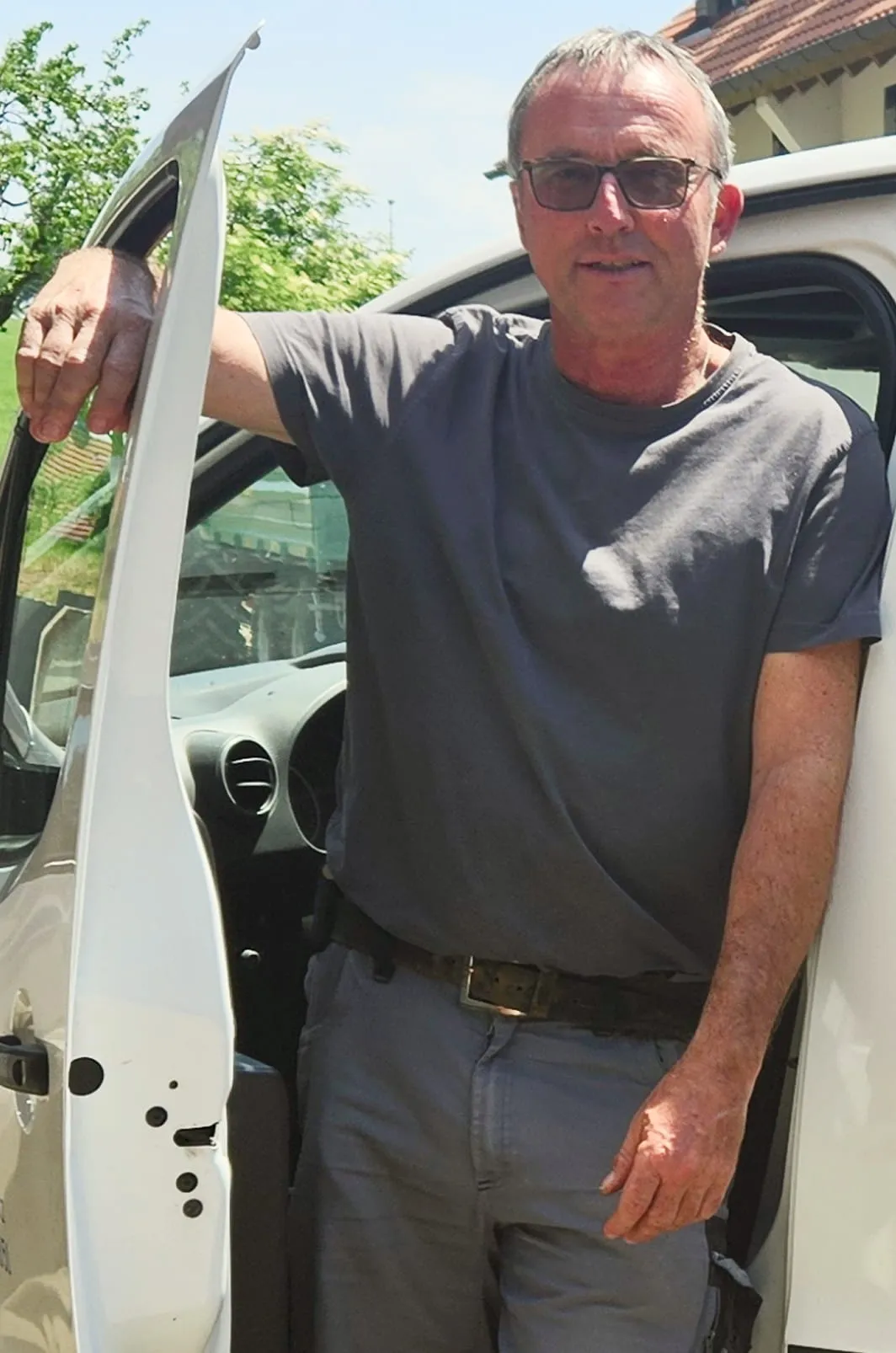Ein Mann mit Brille und grauem Hemd steht an einem sonnigen Tag draußen an einer offenen Autotür.