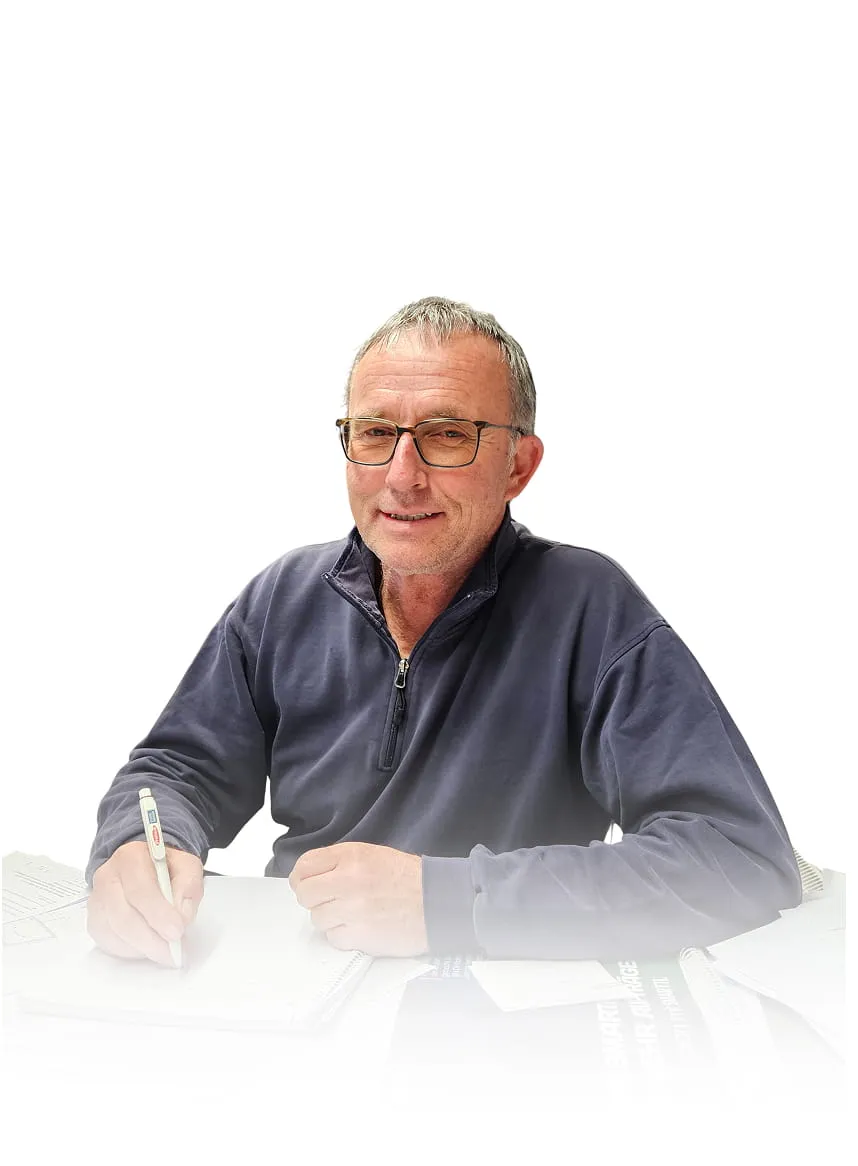 Ein Mann mit Brille und einem dunklen Sweatshirt mit Reißverschluss, der an einem Schreibtisch mit einem Stift schreibt.