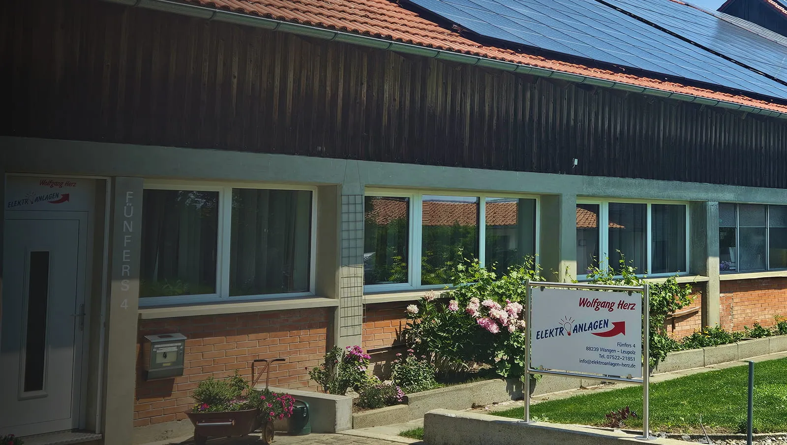Gebäudefassade mit Solarpanelen auf dem Dach, Garten mit Blumen, und ein Schild für Wolfgang Herz Elektroanlagen.