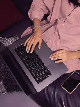 Person in pink shirt typing on a laptop with a smartphone beside them on dark furniture.