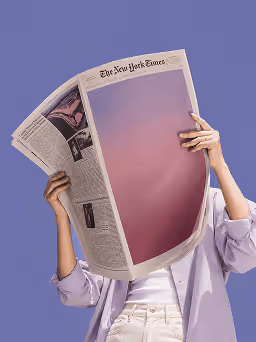 Person in light purple shirt holding up a New York Times newspaper with a blank pink-purple gradient page.