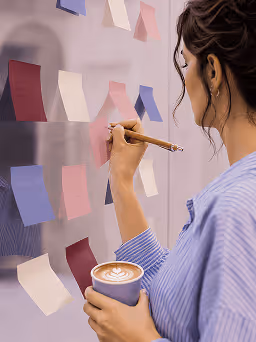 Woman holding a cup and writing on colorful sticky notes on a translucent board.