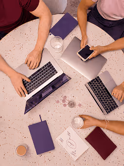 Overhead view of people working with laptops, notebook, smartphone, papers, and drinks on a round table.