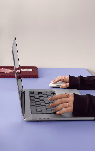 Hands typing on a laptop keyboard and using a mouse on a blue desk with a red notebook nearby.