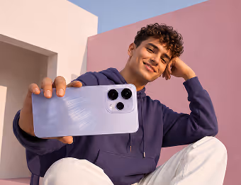 Young man smiling and holding up a smartphone camera toward the viewer against a pink and white background.
