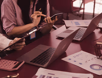 Two people at a desk using tablets and laptops, with charts and a calculator nearby.