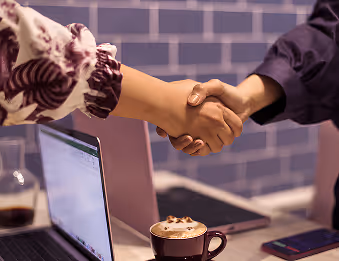 Two people shaking hands above a table with a laptop and a cup of coffee.