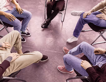 Five people sitting on chairs arranged in a circle with visible legs and hands.