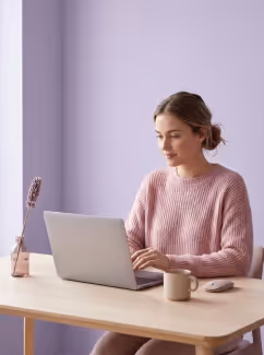Woman in pink sweater working on a laptop at a wooden desk with a mug and a plant.