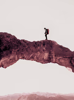 Hiker walking across a natural rock arch with a backpack against a pale sky.
