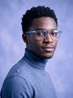 Portrait of a man wearing clear glasses and a gray turtleneck sweater against a blue background.