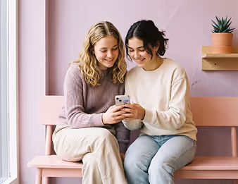 Two young women sitting on a pink bench looking at a smartphone and smiling.
