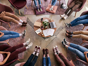 Group of people sitting in a circle with feet towards center around a coffee table with books and plants.