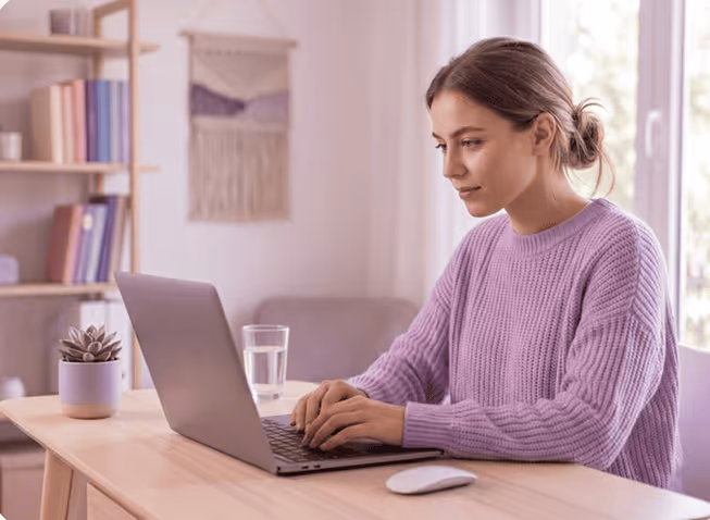 Woman in purple sweater typing on laptop at wooden desk with plant and mouse nearby.