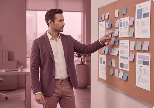 Man in business attire pointing at sticky notes and papers on a bulletin board in an office.