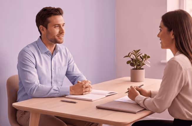 Man and woman with documents having a discussion at a light wooden table with a potted plant.