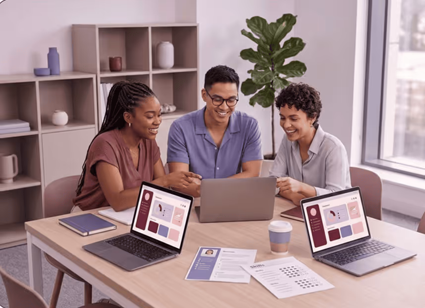 Three colleagues smiling and working together on laptops at a wooden table in a modern office.