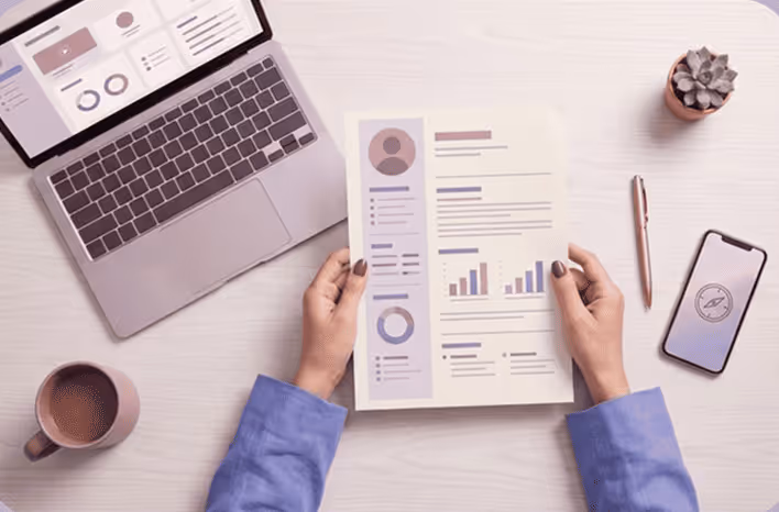 Person holding a resume or report with charts at a desk with laptop, phone, pen, plant, and coffee cup.