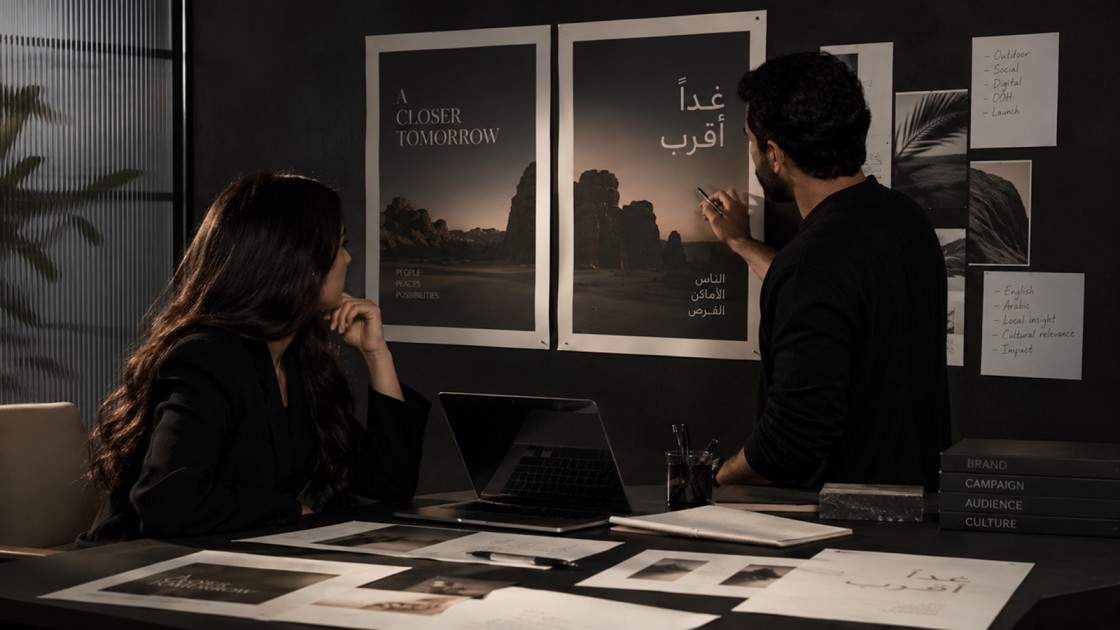Two people reviewing bilingual English and Arabic campaign posters on a dark studio wall, with a laptop and printed layouts on the desk