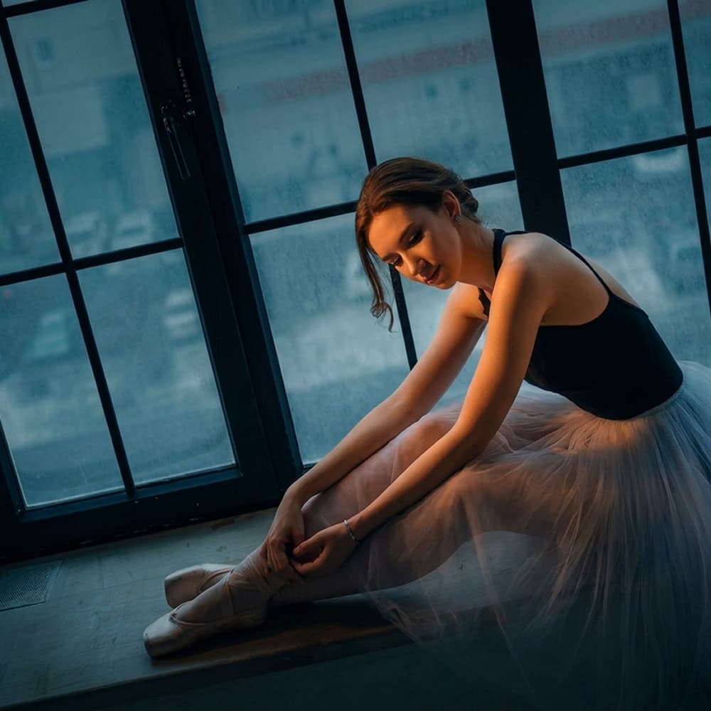 A ballet dancer tying her pointe shoes by a studio window